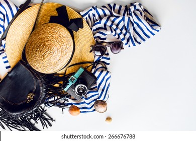 Top view of summer accessories for modern woman on her vacation. Straw hat,  camera, stylish sunglasses, black leather boho bag and striped beach dress on white floor. 