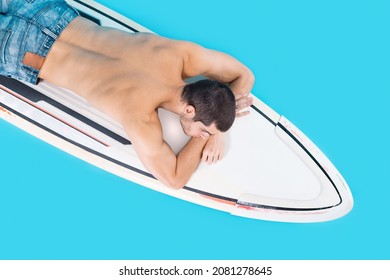 Top View Surfer Laying On Board In Studio On Blue Background, Dreaming, With Naked Torso, Holding Hands Under His Head, Demonstrating His Back.