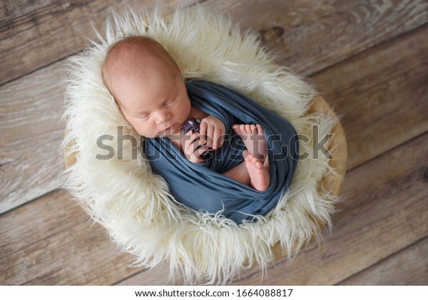top view of sweet newborn boy in blue cover\
keping a small car toy in hands laying in a basket with white fur.\
wooden background