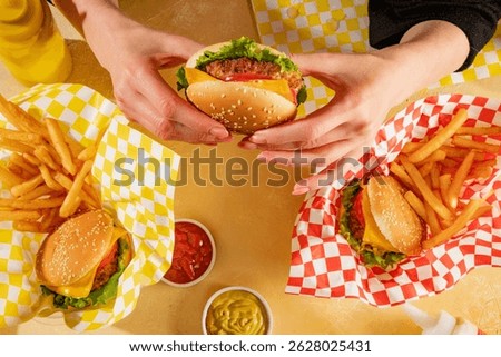 Top view of table composition in yellow tones with fast food - hamburgers in plain wraps, french fries, curry sauce, ketchup and a person holding and eating burger