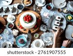 A top view of a table set for dessert with a cake topped with strawberries, pastries, and various condiments. A hand reaches for a plate of treats.