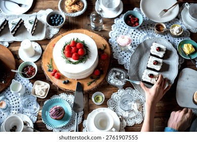 A top view of a table set for dessert with a cake topped with strawberries, pastries, and various condiments. A hand reaches for a plate of treats.