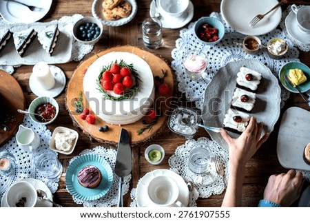 A top view of a table set for dessert with a cake topped with strawberries, pastries, and various condiments. A hand reaches for a plate of treats.
