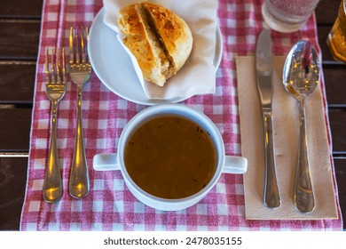 Top view of table setting with bowl of soup, piece of bread on plate, and cutlery arranged on red and white checkered tablecloth. - Powered by Shutterstock
