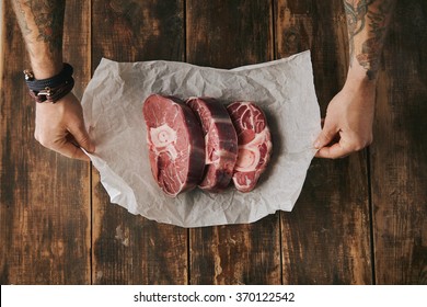 Top View Tattooed Hands Hold White Craft Paper With Three Raw Great Leg Steaks With Bone On It, Everything On Old Wooden Brushed Table