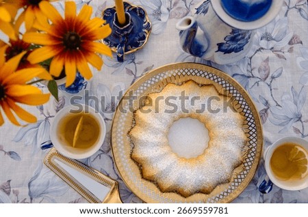 Top view of tea and cake setup on floral tablecloth with yellow flowers and blue porcelain. Summery rural breakfast or tea party atmosphere