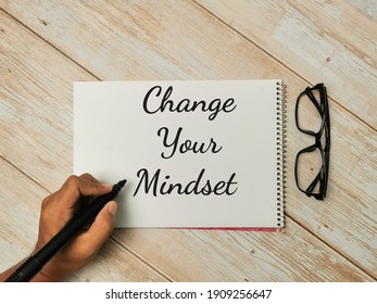 Top View Text Change Your Mindset Written On Notebook With Hand Holding Marker Pen And Eye Glasses On Wooden Table.