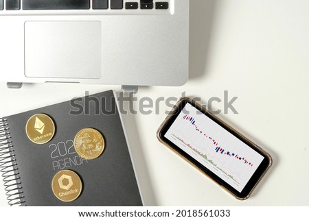 Top view of three different virtual currencies: Bitcoin, Ethereum and Chainlink on a desk next to a laptop and cell phone with investment charts. Concept of new ways to invest in virtual currencies.