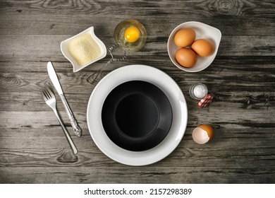 A top view of three red eggs in mug, knife, fork, a sugar plate, and a broken egg yoke in a transparent glass on a gray wooden kitchen table