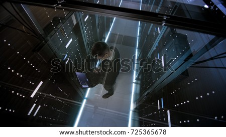 Top View Through the Glass of IT Engineer Working with Laptop in Data Center Full of  Active Rack Servers.
