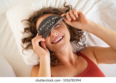 Top view of tired young woman sleeping on bed. Close up face of beautiful sleepy girl wearing funny eye mask while peeping with one eye. High angle view of woman covering her eyes with sleeping mask.