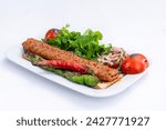 Top view of traditional grilled Adana kebab with cooked spicy shish kebab, pepper, tomato, and fresh greenery on a white plate isolated on white background.