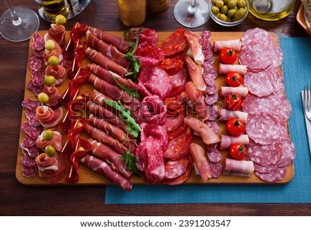 Top view of traditional Spanish meat platter - sliced dry-cured jamon, bacon and sausages on wooden board with olives, tomatoes and greens