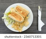Top view of a tuna panini sandwitch in a white plate with crisps, and salad. Knife and Fork wrapped in a tissue on the side.