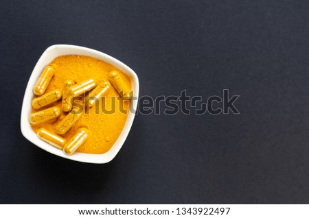 Top view of turmeric powder and capsules in a small white bowl on dark background with copy space. 