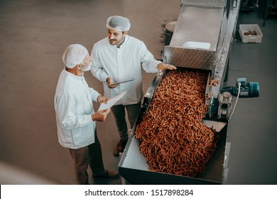 Top view of two Caucasian quality controllers standing next to machine with salty sticks and evaluating quality. Both are dressed in white uniforms and having hairnets. Food plant interior. - Powered by Shutterstock