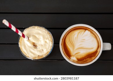 A top view of two coffee drinks placed side by side on a dark wooden slatted table, showing an iced coffee frappe with whipped cream topping and red striped paper straw on the left, and a hot latte with a detailed leaf pattern latte art in a white ceramic cup on the right. The frappe and latte art coffee are photographed from directly above creating a clean flat lay composition on the dark table surface, presenting a classic cafe pairing of cold and hot coffee drinks together