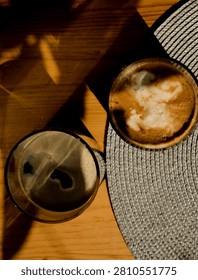 Top view of two coffee drinks on a wooden table with dramatic sunlight and shadows, creating a warm cozy atmosphere. One iced coffee and one milk coffee served on a woven placemat in natural morning light.