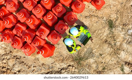 Top view of two construction workers in safety gear using a laptop near a pile of orange traffic cones at a worksite, showcasing teamwork, planning, and efficient use of technology in construction