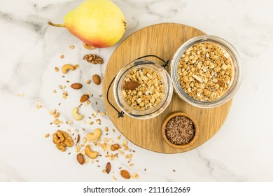 top view of two glass jars with muesli or granola on a wooden board and a marble table with scattered nuts and raisins. proper nutrition