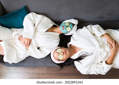 Top view of two relaxed young women stepping out of the shower with a bathrobe and resting on the sofa together with facial masks - Powered by Shutterstock