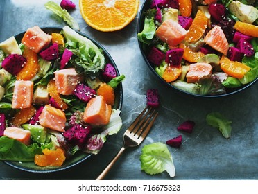Top view of two salmon, orange, purple dragon fruit salad in black bowls on rustic background with a fork