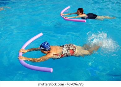 Top View Of Two Senior Women Doing Swimming Exercise With Soft Foam Noodles In Outdoor Swimming Pool.