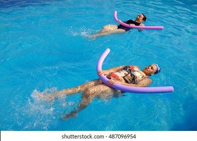 Top View Of Two Senior Women Practicing Backstroke With Soft Foam Noodles In Outdoor Swimming Pool.