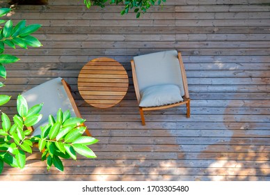 Top View Of Two Stylish Wooden Chairs With Cushions And A Small Wooden Table At A Balcony Of A Room In A Resort, Surrounded By Garden 