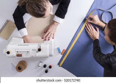 Top View Of Two Women Sewing At One Table. One Is Using Her Sewing Machine. The Second Is Measuring