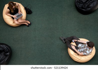 Top view of two young people, man and woman, sitting on bean bags, using electronic devices in public wifi area, working on laptop, typing, using smartphone, texting message, top view - Powered by Shutterstock