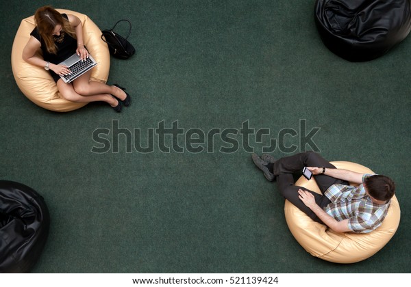 Top\
view of two young people, man and woman, sitting on bean bags,\
using electronic devices in public wifi area, working on laptop,\
typing, using smartphone, texting message, top\
view