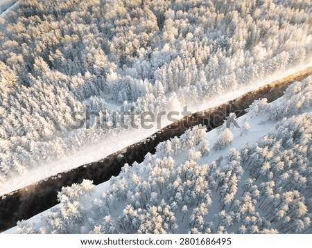 Top View of Unfrozen River Through Snowy Fores. Top down aerial view of a dark unfrozen river cutting through a snow covered forest, with light mist above the water during severe winter frost.