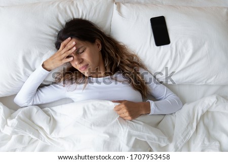 Top view unhappy woman feeling headache after sudden awakening by phone call, message signal or alarm in early morning, exhausted young female suffering from insomnia or migraine, lying in bed