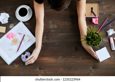 Top view of unrecognizable woman clearing up space on wooden desk, putting away stationery, papers, plant, food and other stuff