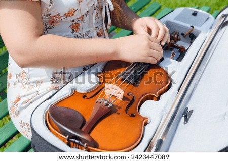 top view unrecognizable young artist woman wearing dress and sitting on wooden bench in park on street unzipping violin from case to take it out. copy space.