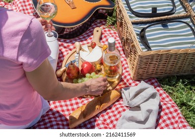 Top View Of Unrecognizable Young Woman In White Pants Outside Having Picnic, Eating And Playing Guitar. Summer Fun And Leisure. View From Behind