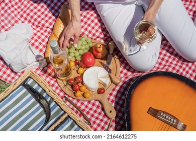 Top View Of Unrecognizable Young Woman In White Pants Outside Having Picnic, Eating And Playing Guitar. Summer Fun And Leisure