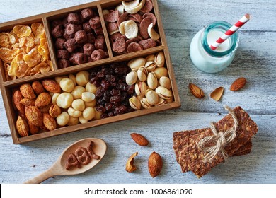 Top View Of Variety Of Nuts, Cereal And Raisin In Wooden Box, Homemade Cereal Bar And Almond Milk Over White Table. Healthy Breakfast For Kids. 