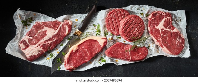 Top view of various uncooked ribeye and sirloin beef steaks arranged with burger cutlets and sharp knife seasoned with herbs - Powered by Shutterstock