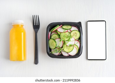 Top View Of Vegetarian Salad Made Of Sliced Cucumber And Radish Served In Black Plastic Bowl With Bottle Of Orange Fruit Juice And Telephone With Copy Space Or Mock Up On White Wooden Background