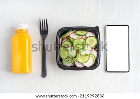 Top view of vegetarian salad made of sliced cucumber and radish served in black plastic bowl with bottle of orange fruit juice and telephone with copy space or mock up on white wooden background