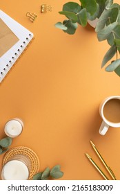 Top view vertical photo of workplace cup of coffee note pads gold pens binder clips candles on rattan serving mat and vase with eucalyptus on isolated orange background with copyspace in the middle