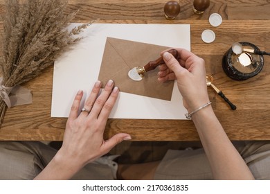 Top view of well-kept hands of unrecognizable woman making white wax seal with stamp on brown greeting envelope.