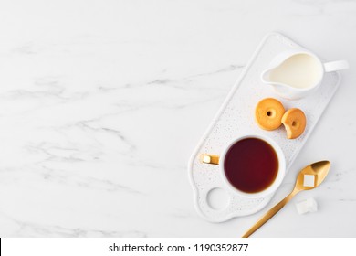 Top View Of White Cup Of Tea, Biscuits And Milk Jug On Serving Plate, Golden Spoon With Sugar Cubes On White Marble Background. Flat Lay With Copy Space.