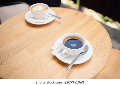 Top View Of White Cups Of Aromatic Coffee Americano And Cappuccino On A Wooden Table, In The Summer Terrace Of A Cafeteria. Food And Drink. Copy Advertising Space. Still Life