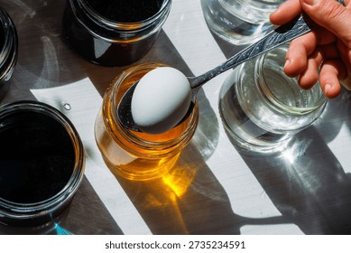 Top view of a white egg held on a spoon above a jar of yellow food dye, ready to dip, surrounded by clear water jars in bright sunlight.