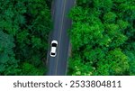 Top view of a white electric car driving  dark green forest road, which is an elevated road that surrounds natural forest. Concept of using electric cars to protect the environment and transportation.