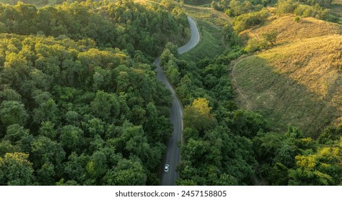 Top view of a white electric car driving  dark green forest road, which is an elevated road that surrounds natural forest. Concept of using electric cars to protect the environment and transportation.