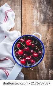 Top View Of A White Metal Colander With Cherries On A Rustic Wooden Table With A White Kitchen Rag, With Copy Space